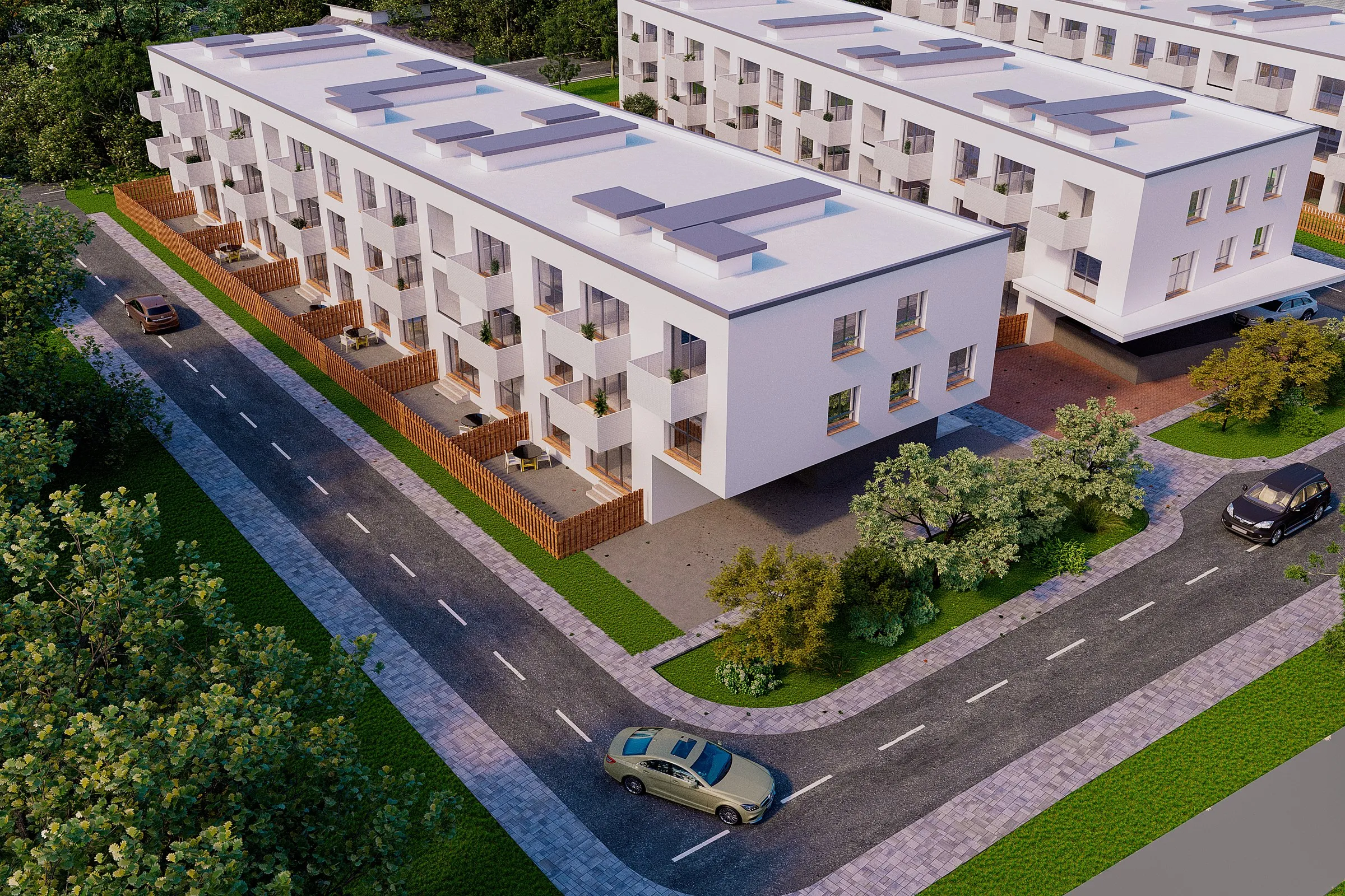 Aerial view of white three-storey residential blocks with balconies and fenced ground-floor terraces, beside a tree-lined street with parked and passing cars